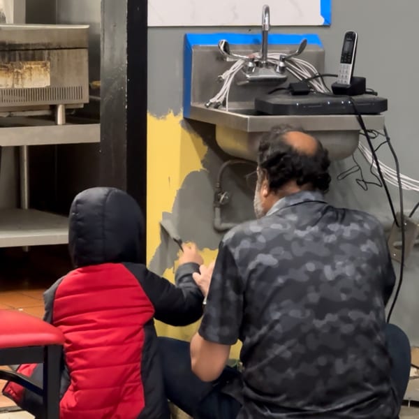 A man and child painting a yellow wall gray next to a sink in a deli kitchen.