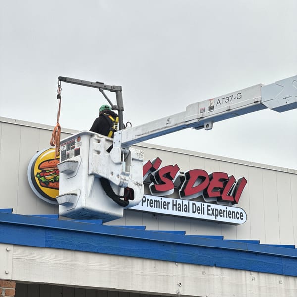 Worker in a lift bucket installing a sign for Jay's Deli, featuring a burger logo.