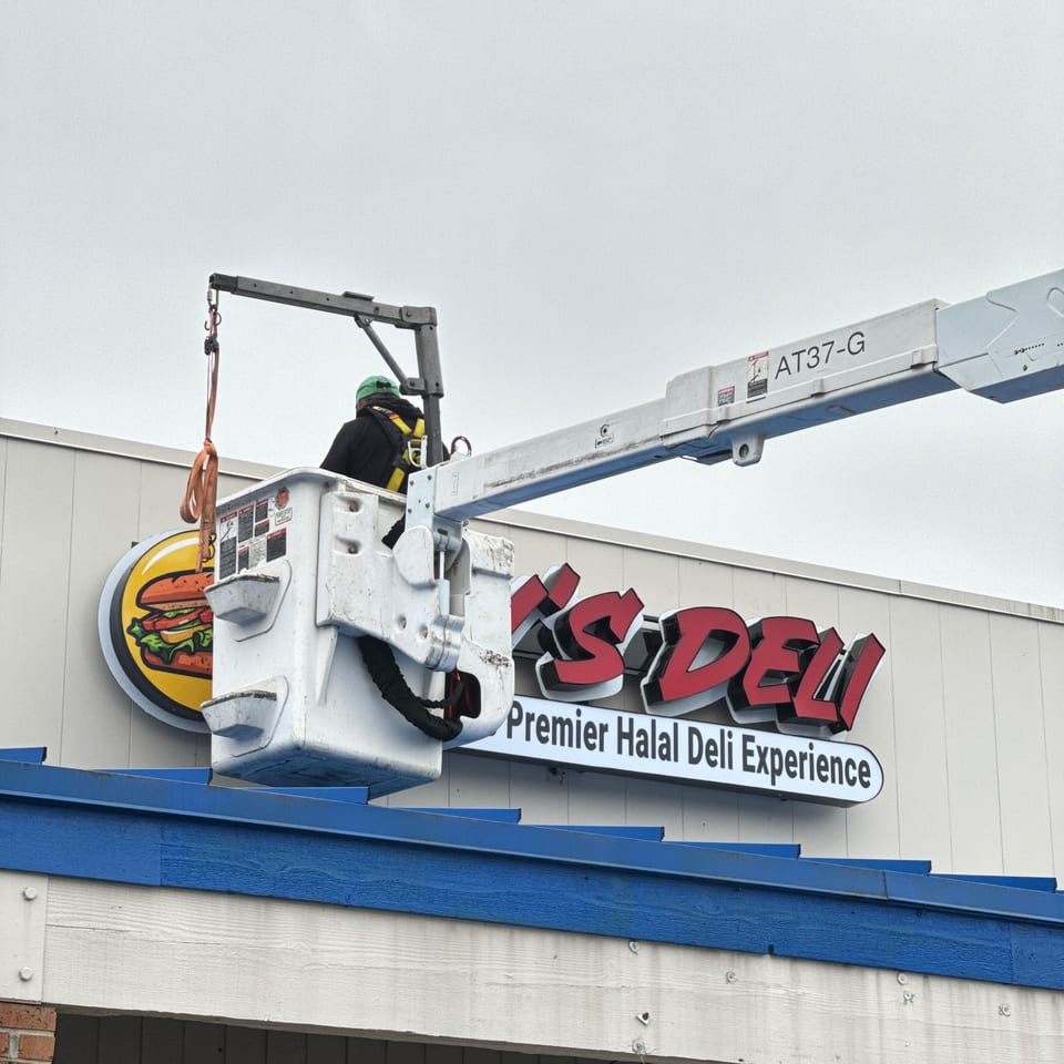 Worker in a lift bucket installing a sign for Jay's Deli, featuring a burger logo.