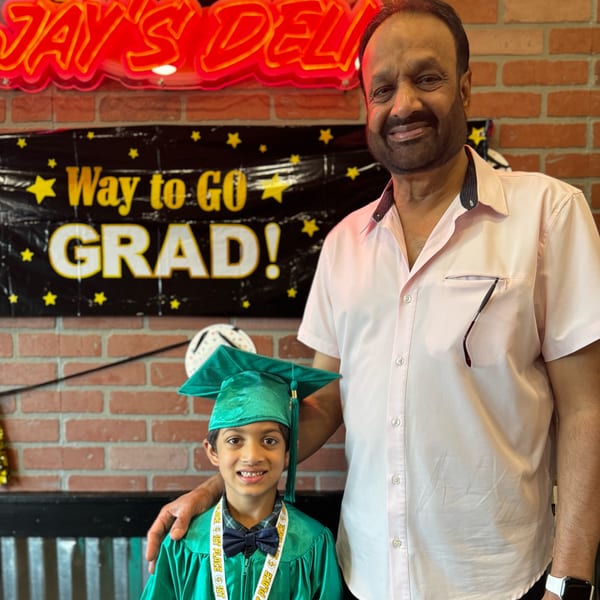 A man and a boy in a green graduation cap and gown smile in Jay's Deli.