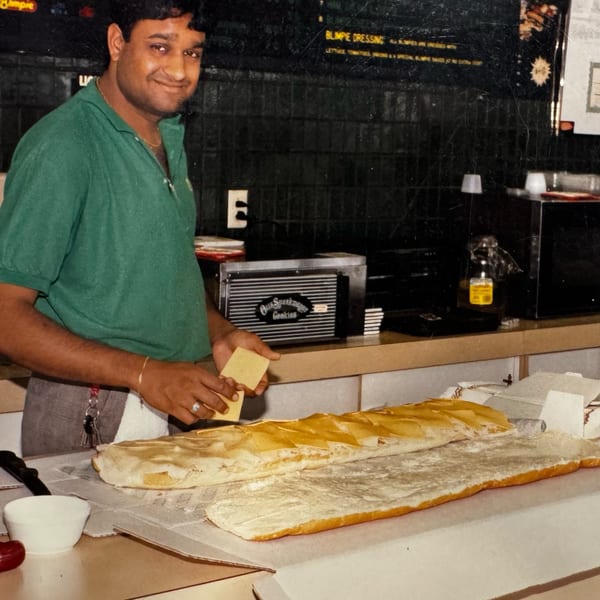 Jay's Deli owner smiles while preparing a large deli sandwich with cheese on a long baguette.
