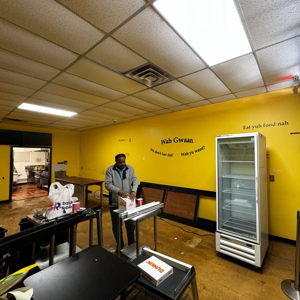 A man stands in a brightly lit deli with yellow walls, packing Dunkin' boxes.