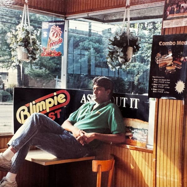 Man in a green shirt and jeans relaxing inside Jay's Deli, with Blimpie branding visible.