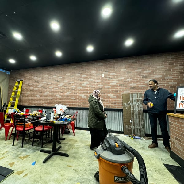 Two people standing in a deli under construction, with exposed brick walls and a vacuum cleaner.