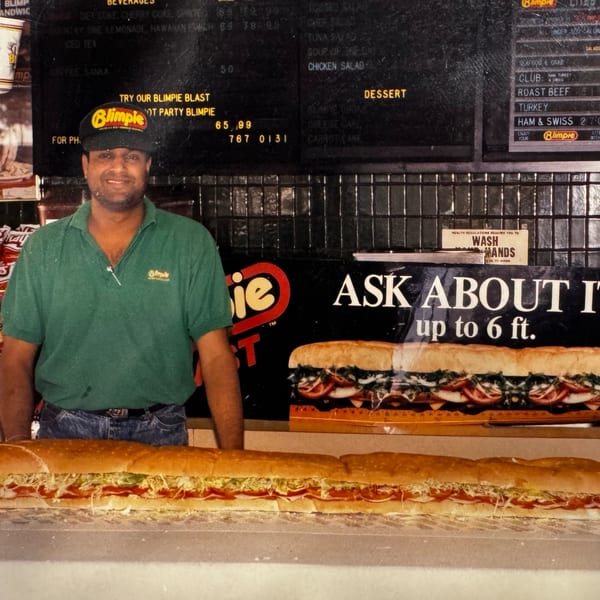 A smiling deli worker in a green shirt and Blimpie hat stands behind a long sub sandwich.